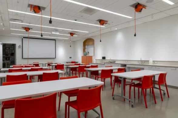 A classroom featuring white tables and red chairs as well as a monitor for video.