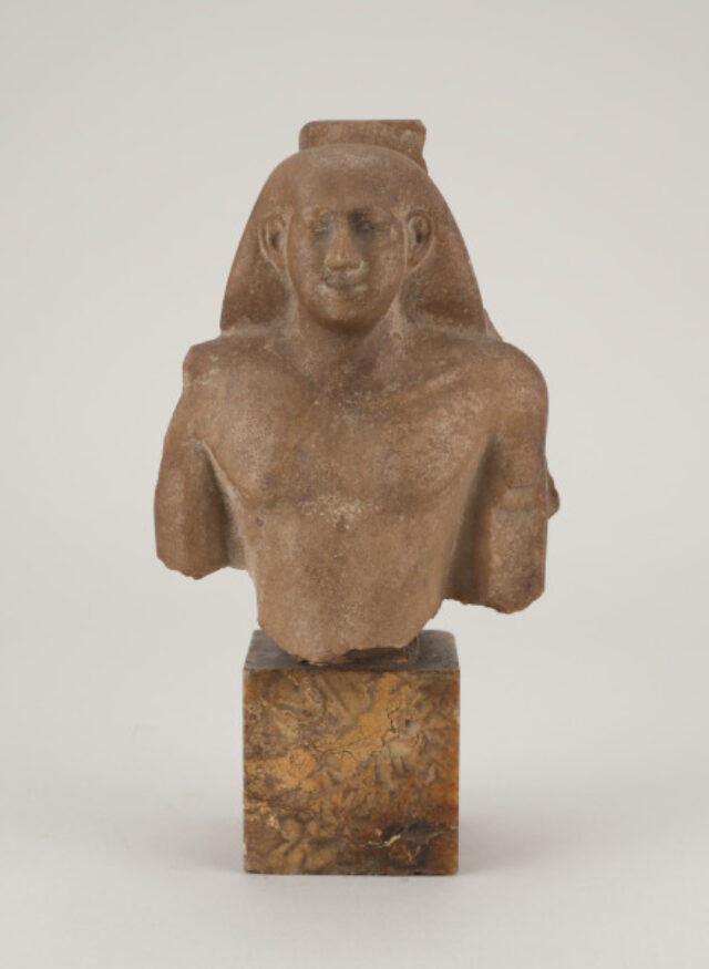 A small stone bust of an ancient Egyptian figure with a headdress typical of Egyptian royalty mounted on a cube of marble-like stone.