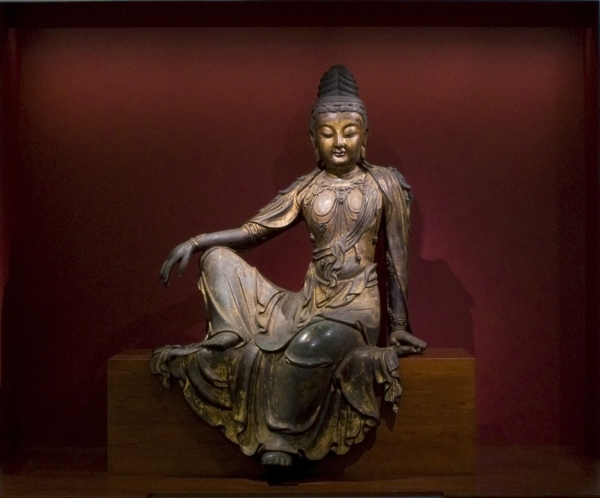 A life-size seated statue of a Boddhisatva figure in a relaxed pose, with one arm resting on its knee and its clothing draped over the edge of a platform. The figure is adorned in many necklaces and bracelets.